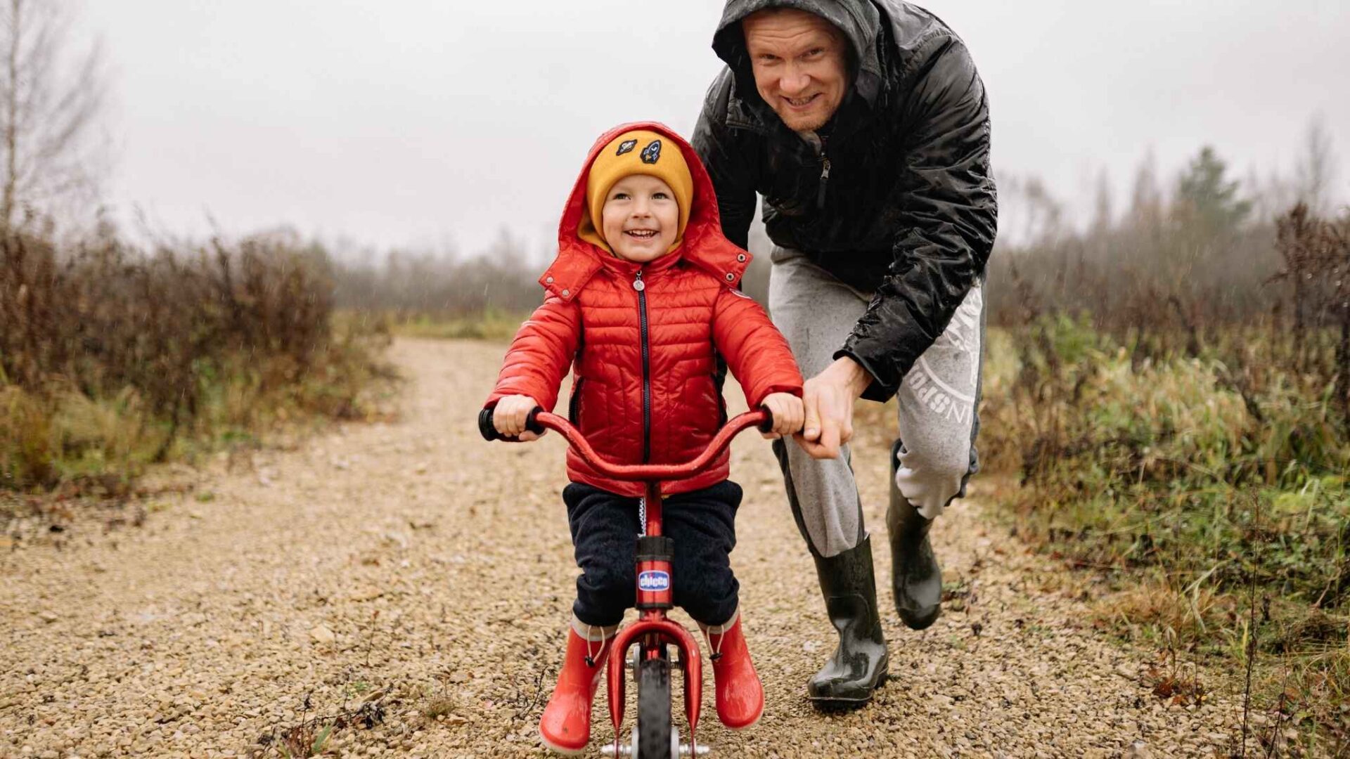 Niño pequeño sonriente montando bicicleta mientras un adulto lo ayuda en un camino de campo al aire libre